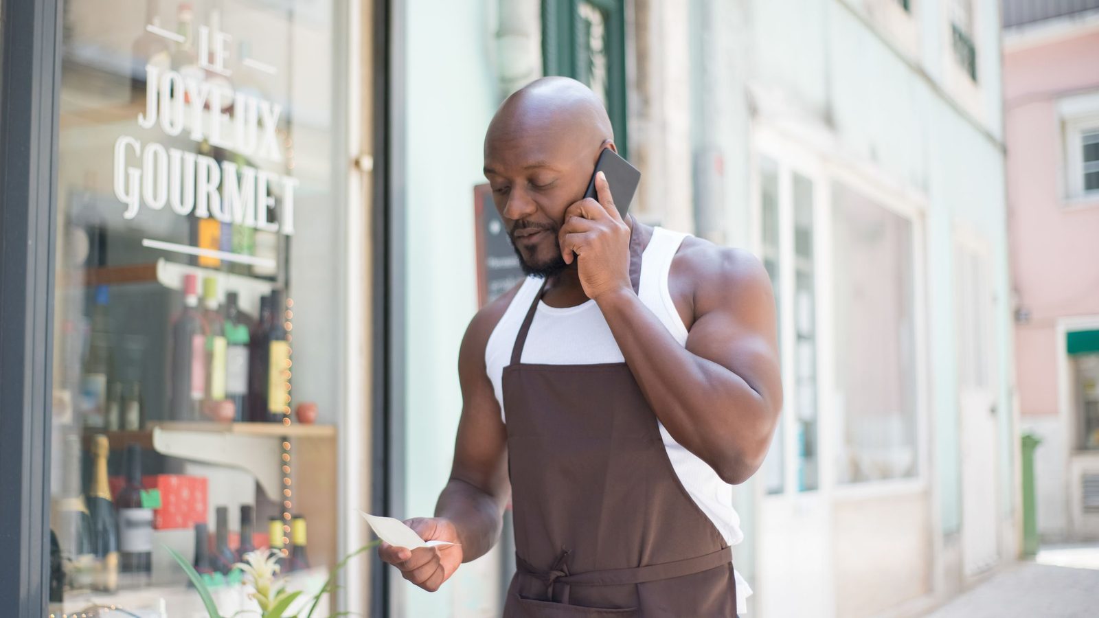 Commerçant en tablier, au téléphone devant sa boutique de quartier « Le Joyeux Gourmet »